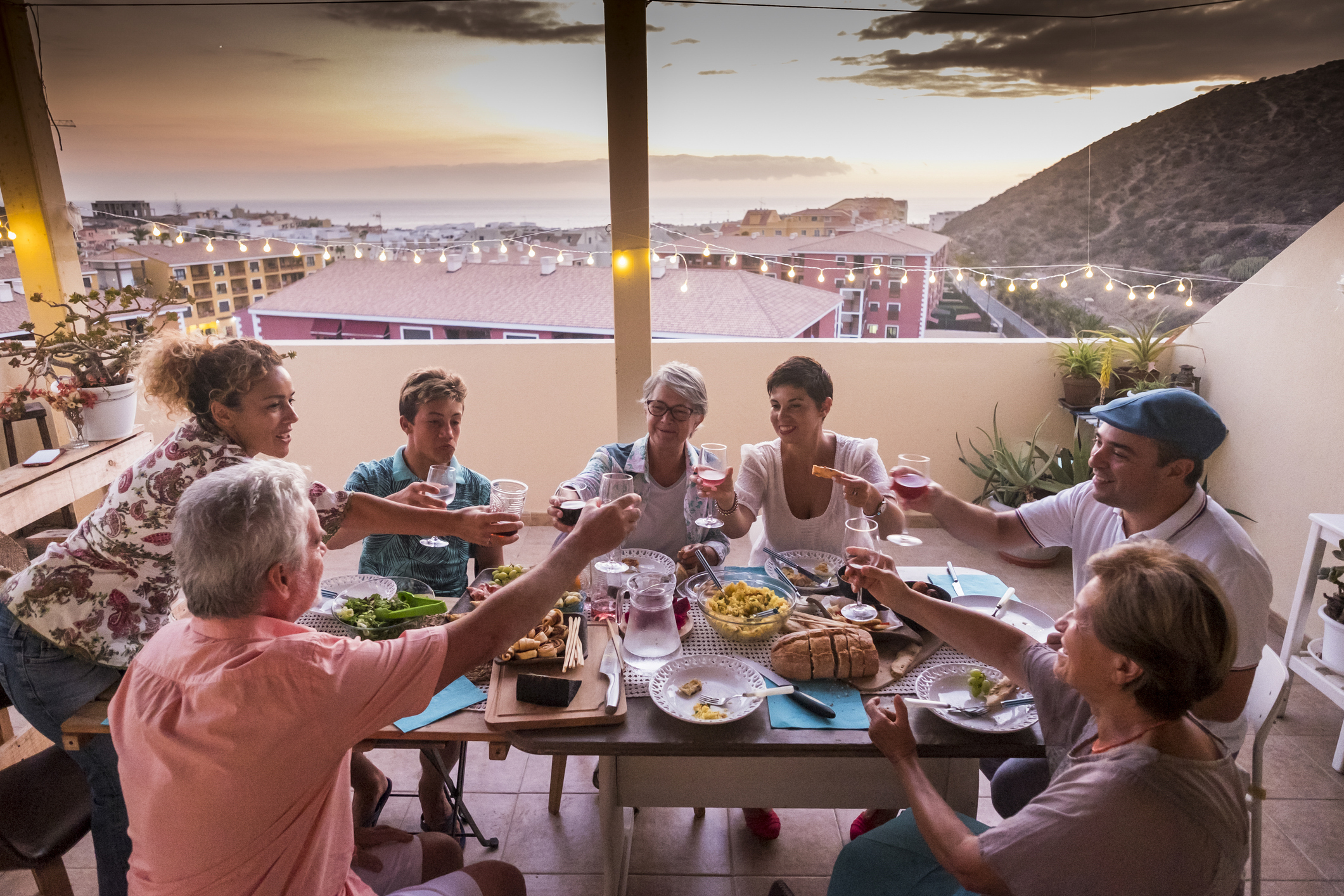 group of friends and parents at home dining together and clinking glasses with beverages. everybody smile and have fun celebrating in friendship. happy caucasian people on the terrace at home with buildings and sea view  during sunset